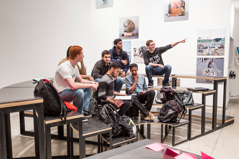 People sit on Blackwell's sculpture. One visitor points to something outside of the frame. Mary Mattingly's prints are installed on the wall behind the sculpture.