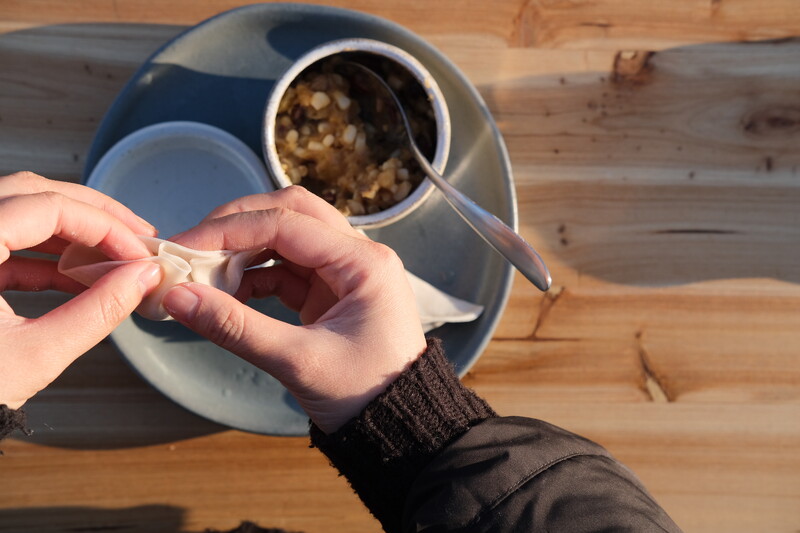 A person pinches a dumpling wrapper into pleats to close it. Underneath their hands, a bowl of dumpling filling sits on a wooden table.