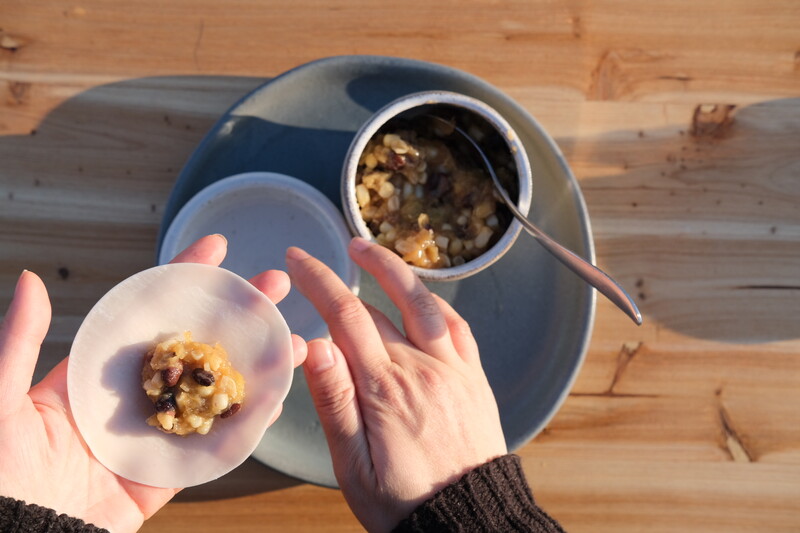 Photographed from above, a person is shown in the process of making dumplings. In their left hand is a dumpling wrapper with a mixture of corn, beans, and squash in the centre. Underneath them, a bowl of the dumpling filling sits on a wooden table.