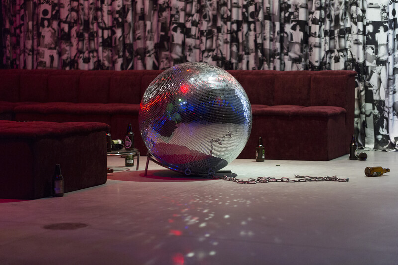 A large disco ball centered on the floor of a dimly lit room in front of a red velvet couch and coffee table. Black and white photographs appear on the wall behind the couch, and empty beer bottles are visible on the floor around the couch.