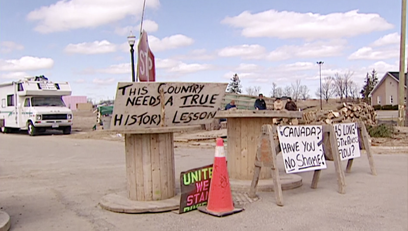 A grainy film still image of the blockade in Caledonia shows the large wooden wire spools and trestles used to block the road. The objects are also being used to hold protest signs that read, “THIS COUNTRY NEEDS A TRUE HISTORY LESSON”, “CANADA? HAVE YOU NO SHAME?”, and “UNITED WE STAND”, held up and partially covered by an orange pylon. The residential development has stop signs and streetlights, some figures are standing in the background with an RV parked nearby.