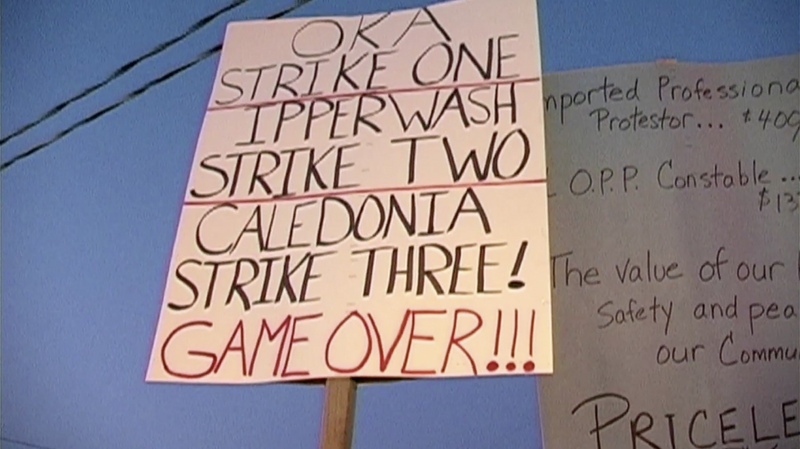 A grainy film still image looks up at two protest posters, one reads “OKA STRIKE ONE, IPPERWASH STRIKE TWO, CALEDONIA STRIKE THREE, GAME OVER!!!”, the second is partially cut out of frame and illegible.&nbsp;